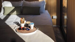A modern hotel room with a large bed, a bedside table with breakfast items, and a view through a large window.