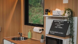 A compact kitchen with a wooden countertop, a sink, a black dishwasher, a microwave, and a sign welcoming Matt to Little Oakey Acres Tiny Home Escape.