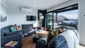 A modern living room with a grey sofa, a round coffee table, and a large window offering a mountain view. - Tiro Nui in the Heart of Wanaka (Wānaka)