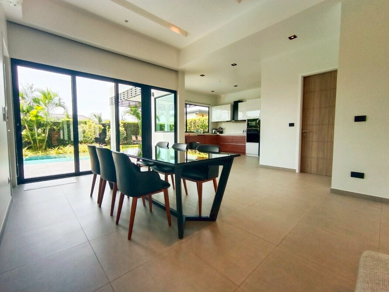 A spacious dining area with a glass table and chairs, a kitchen in the background, and a sliding glass door leading to a garden.