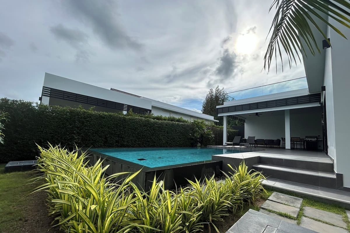 Un área de piscina moderna al aire libre con una piscina rectangular, rodeada de exuberante follaje y un césped bien cuidado.