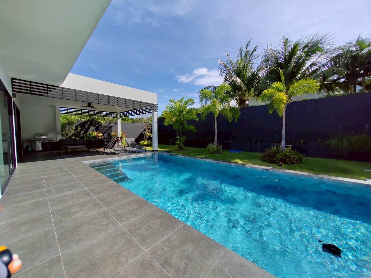 A modern outdoor swimming pool with a covered seating area and a view of the surrounding greenery.