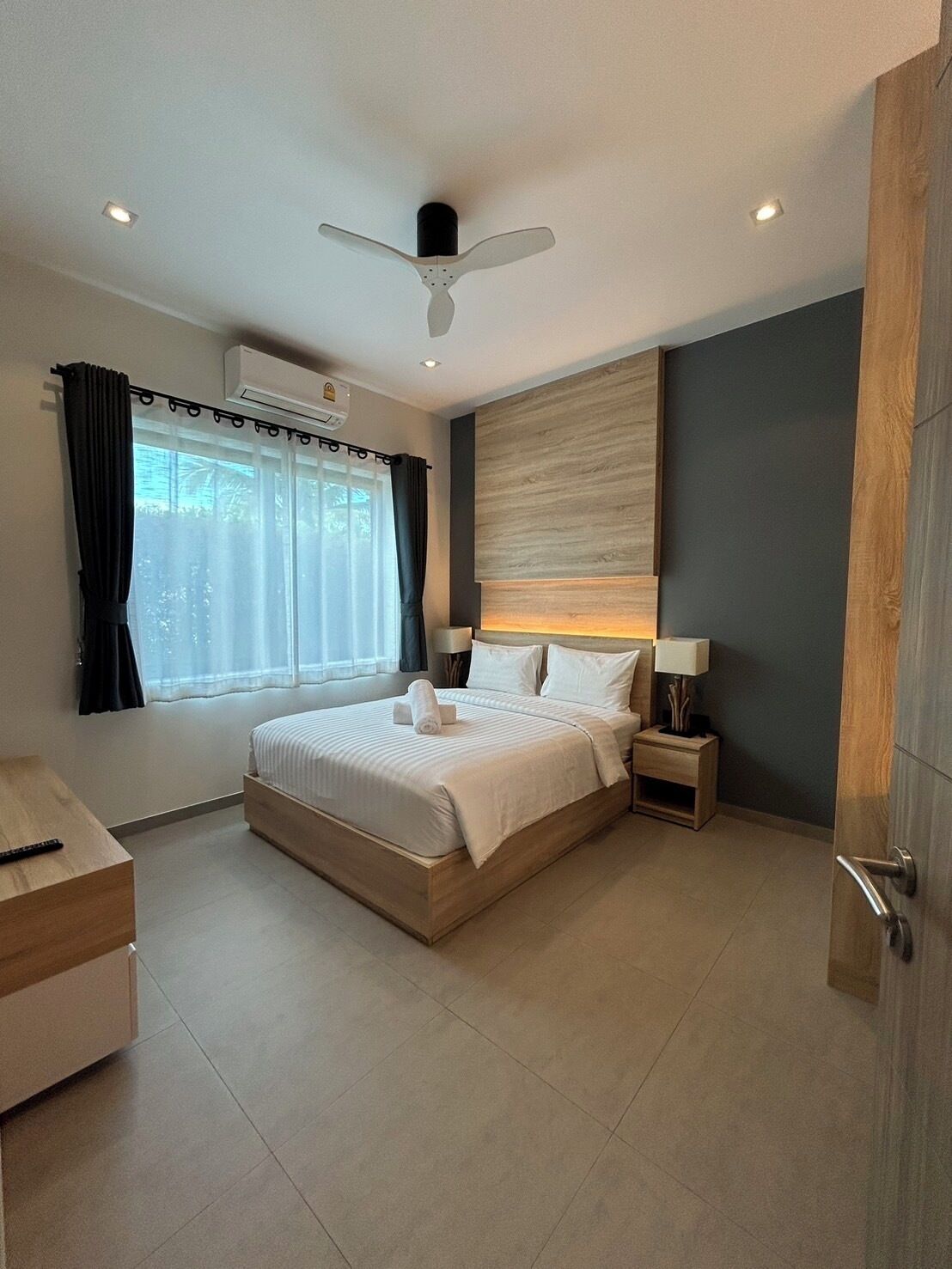 Une chambre à coucher moderne avec un grand lit, un ventilateur de plafond, un appareil de climatisation, une table de chevet avec une lampe et une vue sur la verdure par la fenêtre.