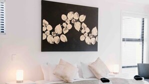 A bedroom with a bed, white nightstands, a bedside lamp, a large wall art piece, and a window with blinds. - Nelson Central Town House (Nelson)