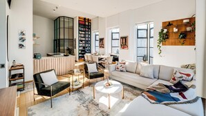 A spacious living area with a large sofa, a coffee table, and a dining table. There is a bookshelf filled with books and a kitchen area visible in the background. - Contemporary 2bed in Art Deco Heritage Site Elevea (Cape Town)