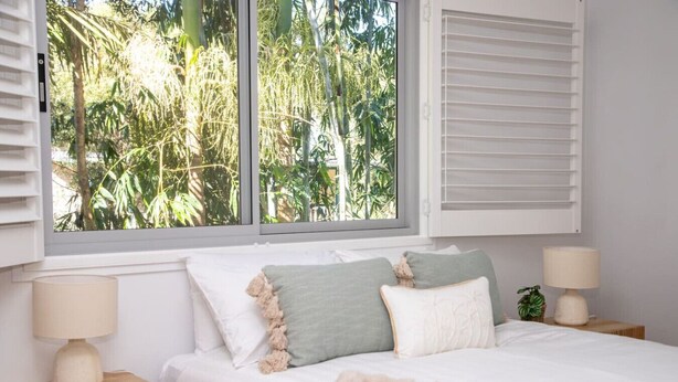 A bedroom with a large window, a bed with white bedding and multiple pillows, two bedside tables with lamps, and a view of greenery outside.