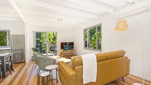 A modern living room with a sofa, a dining area with chairs, a TV, and a large window with a view of greenery.