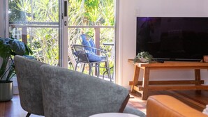 A modern living room with a large TV, a grey sofa, a wooden coffee table, and a balcony with plants and a chair.