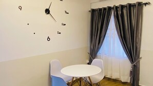 Una habitació amb un rellotge de paret, una taula rodona amb dues cadires, una finestra amb cortines i un aparell d'aire condicionat.
