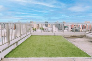 Una terraza en la azotea con césped, barandilla metálica y vistas a la ciudad.