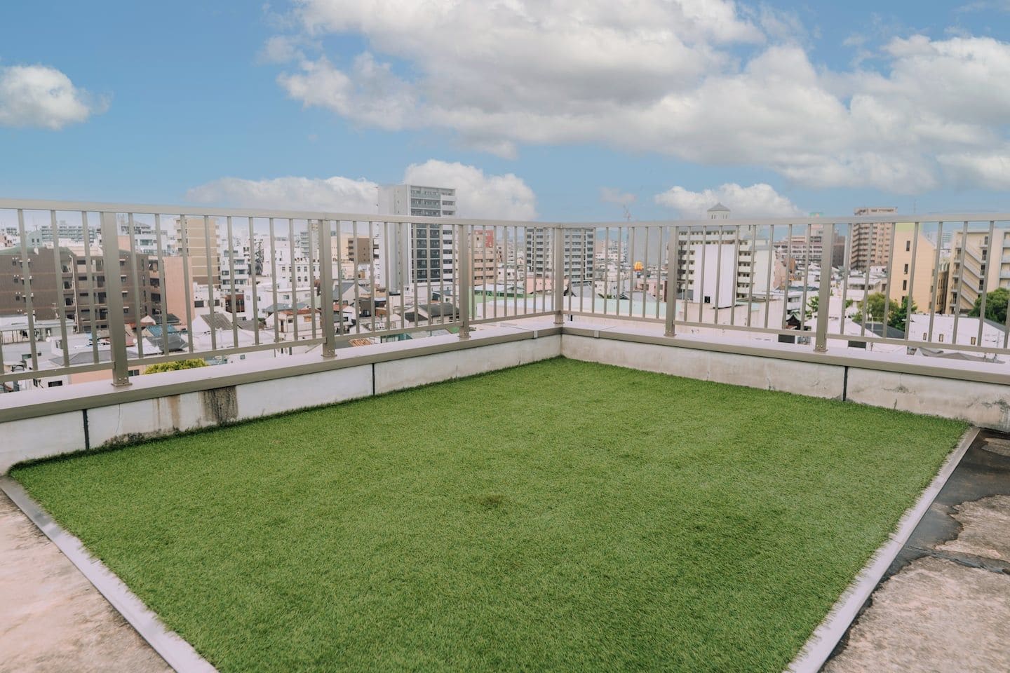 Terraza con área verde delimitada por una barandilla metálica, con vistas a un paisaje urbano y diversos edificios.