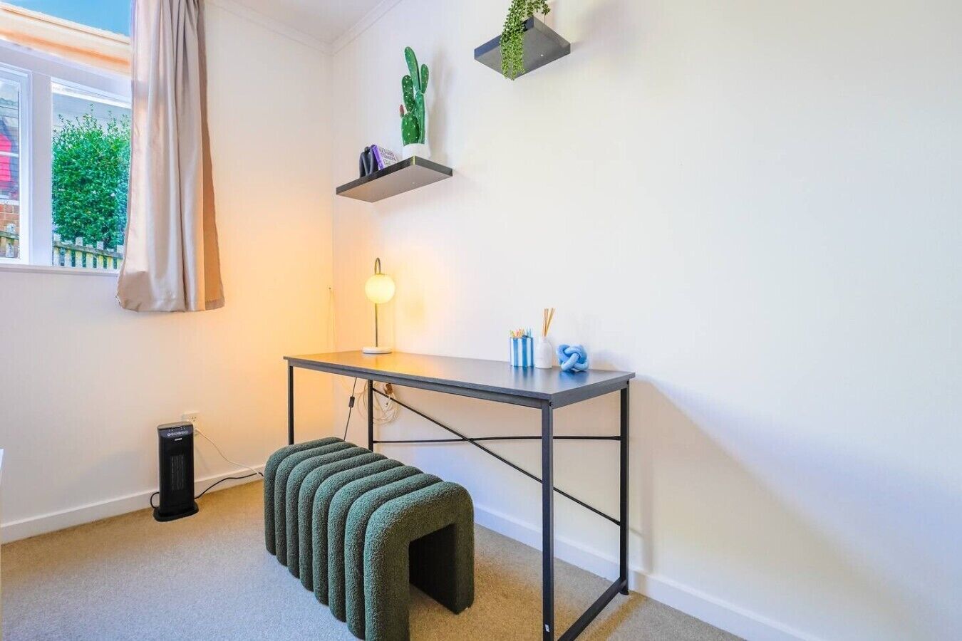 Et værelse med skrivebord, en lille bænk, en lampe og to hylder med planter.