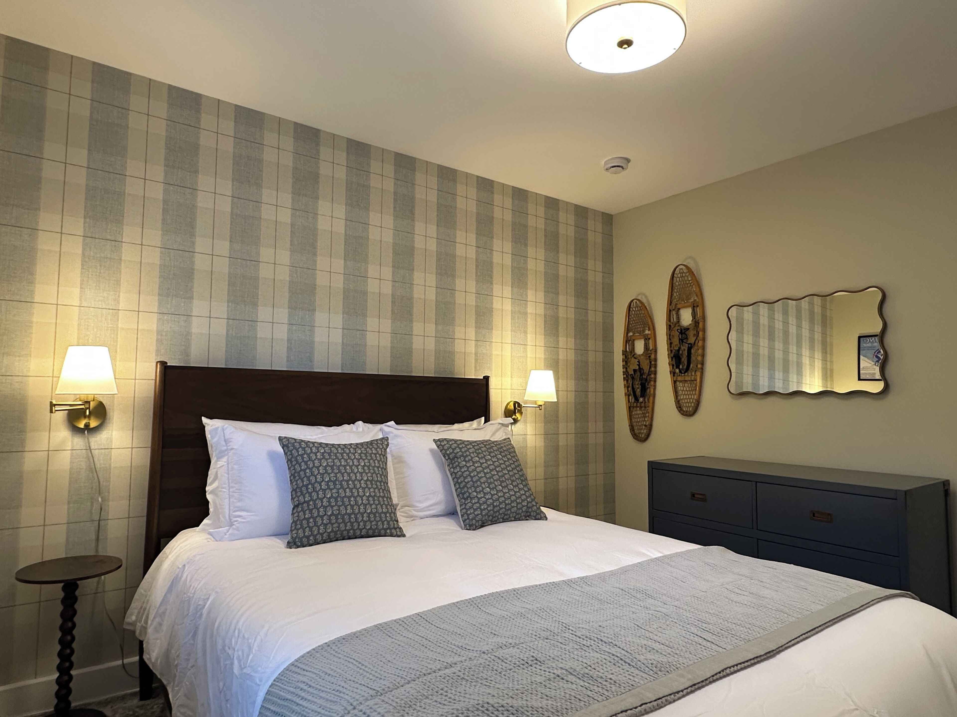 Un lit bien fait, avec une couette blanche et des coussins à motifs, deux lampes fixées au mur, un miroir décoratif et une petite table de chevet.