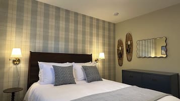 Un lit bien fait, avec une couette blanche et des coussins à motifs, deux lampes fixées au mur, un miroir décoratif et une petite table de chevet.