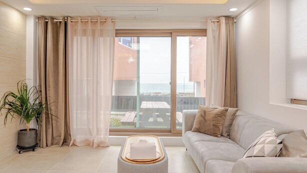 Een moderne woonkamer met een bank, voetenbankje en uitzicht op het groen door grote ramen.