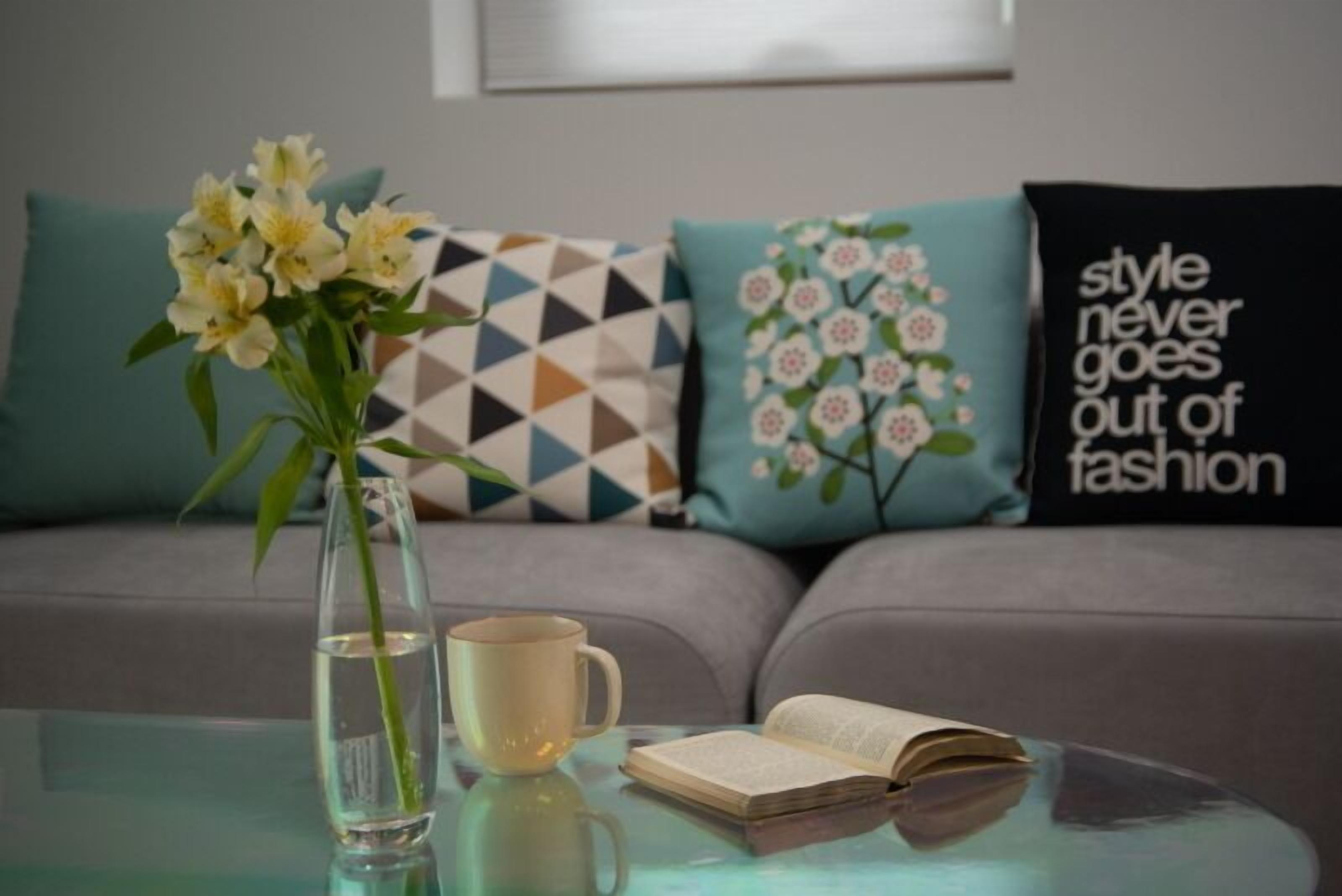 A modern living room with a glass coffee table, a vase with flowers, a book, and a mug.