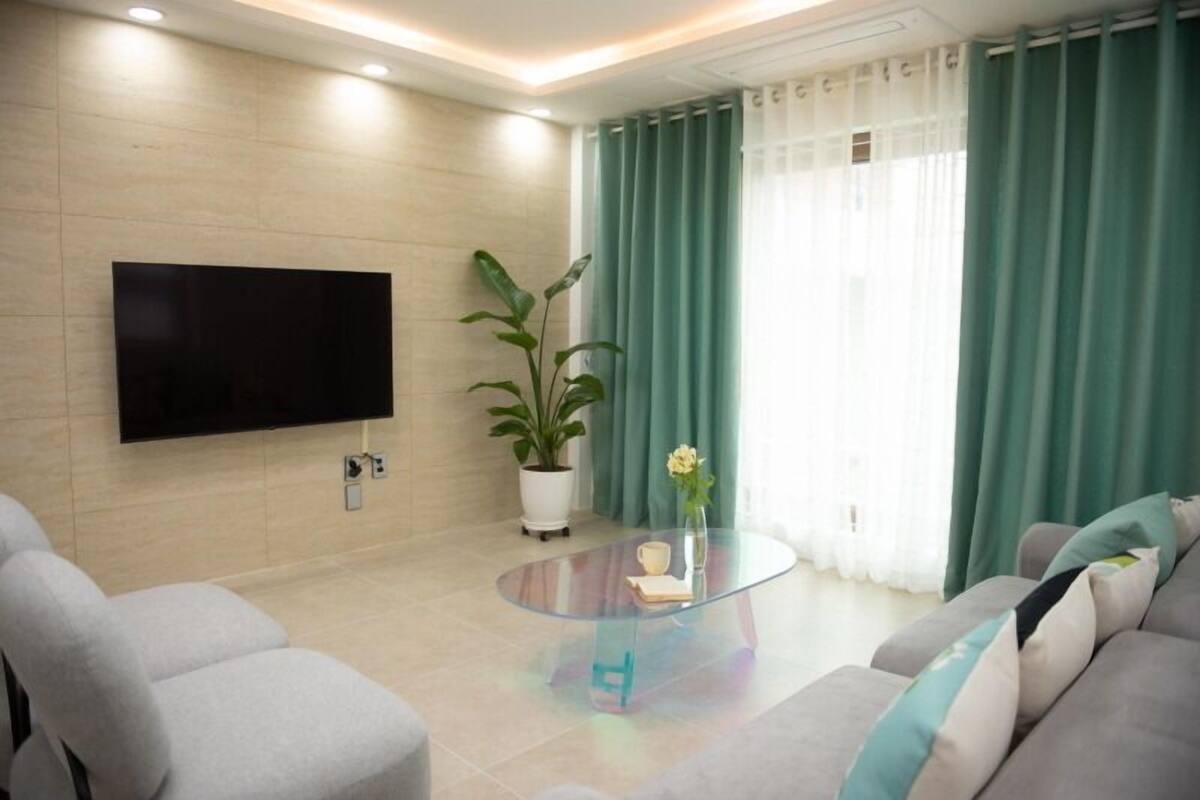 Ein modernes Wohnzimmer mit einem an der Wand befestigten Flachbildfernseher, einem Glastisch, einer Topfpflanze und einer Couch mit Kissen.