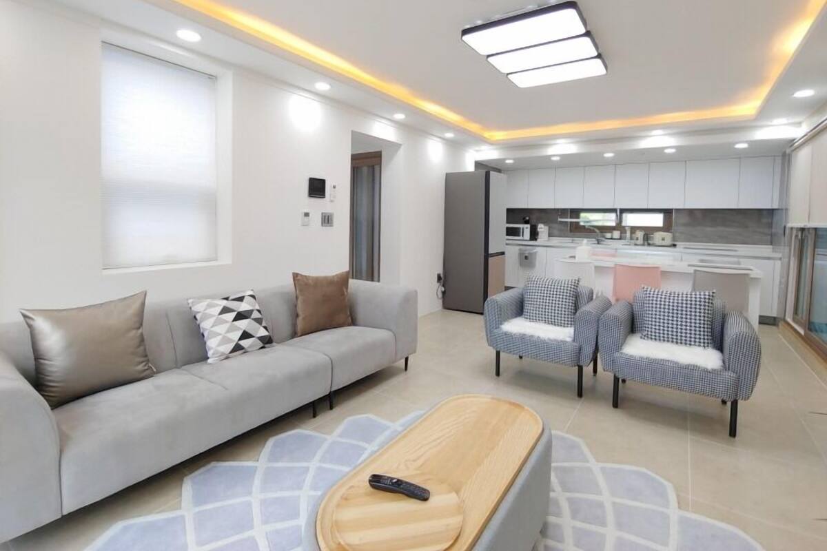 Ein modernes Wohnzimmer mit einer Couch, Sesseln und einem Couchtisch. Im Hintergrund ist eine Küche erkennbar.