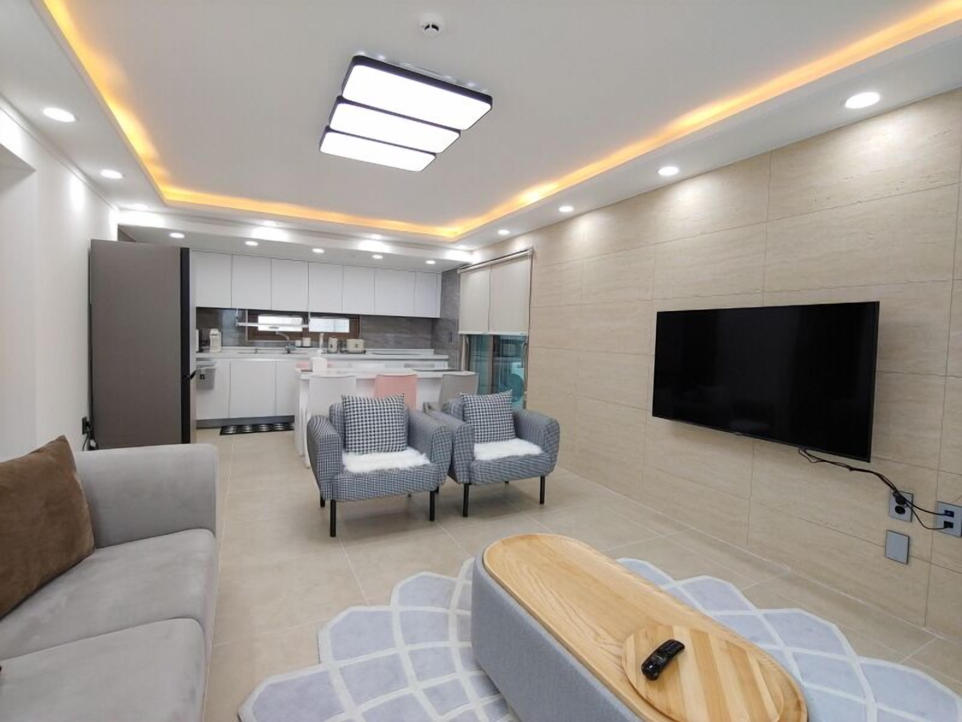 A modern living room with a sofa, two armchairs, a TV, and a kitchen in the background.