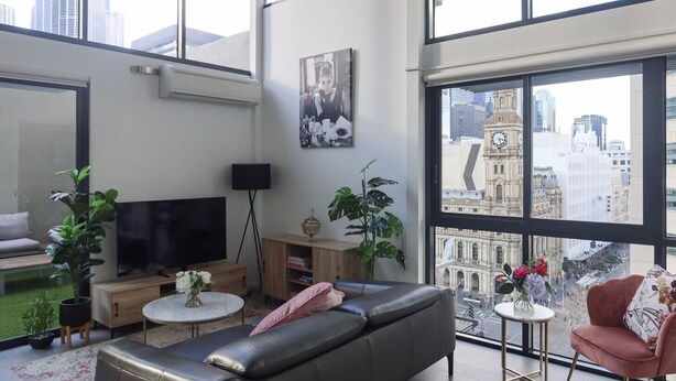 A modern living room with a sofa, a TV, a coffee table, and a view of a cityscape through large windows. - Rare Loft Style Penthouse Bourke St Mall + Parking (Melbourne)
