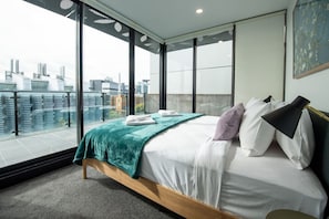 A modern bedroom with a large window, a bed with white linens, a teal throw blanket, and a city view.