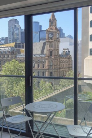 Un balcon avec deux chaises blanches et une petite table, d’où l’on peut admirer un bâtiment historique doté d’une tour horloge, ainsi que des gratte-ciel modernes en arrière-plan.