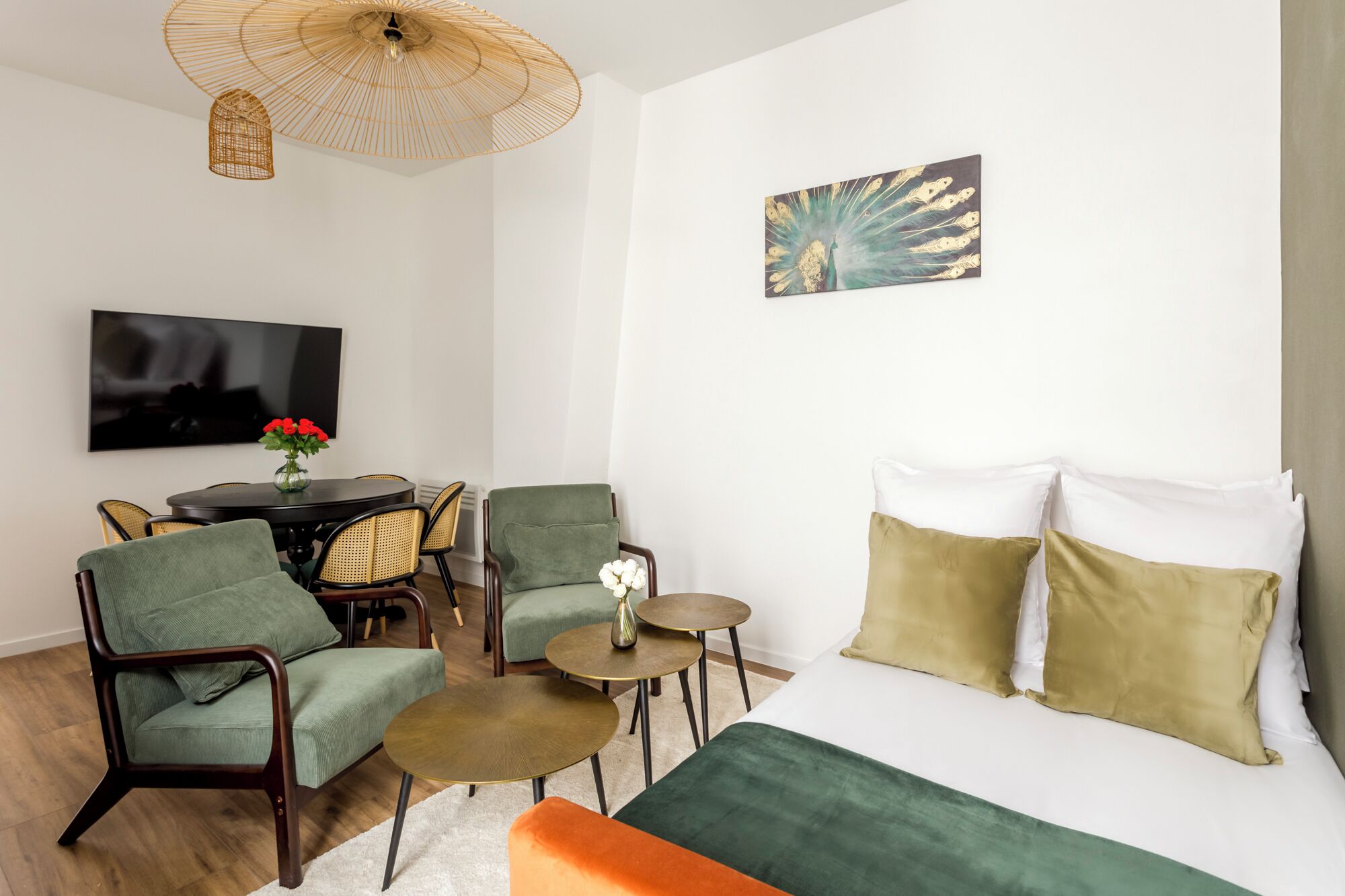 A modern hotel room with a bed, green armchairs, a small table, a television, and a decorative wall piece.
