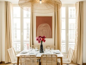 A dining area with a table set for four, surrounded by wicker chairs, a large woven umbrella light fixture, and a vase of flowers. - Family Suites Caumartin I-II (Paris)