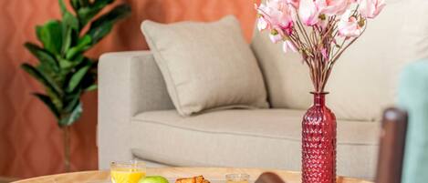 A round wooden table with a plate of croissants, a glass of orange juice, and a glass of coffee. A red vase with pink flowers is on the table.