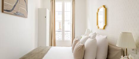 A hotel room with a bed, a bedside table with a lamp, a mirror, and a window with curtains.