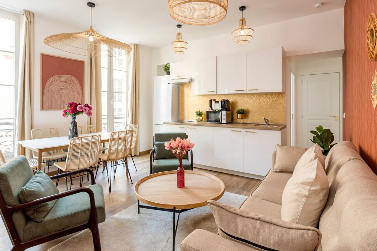 Ein modernes Wohnzimmer mit Sofa, Sesseln, einem Esstisch und einer Küchenzeile mit Schränken und Geräten.