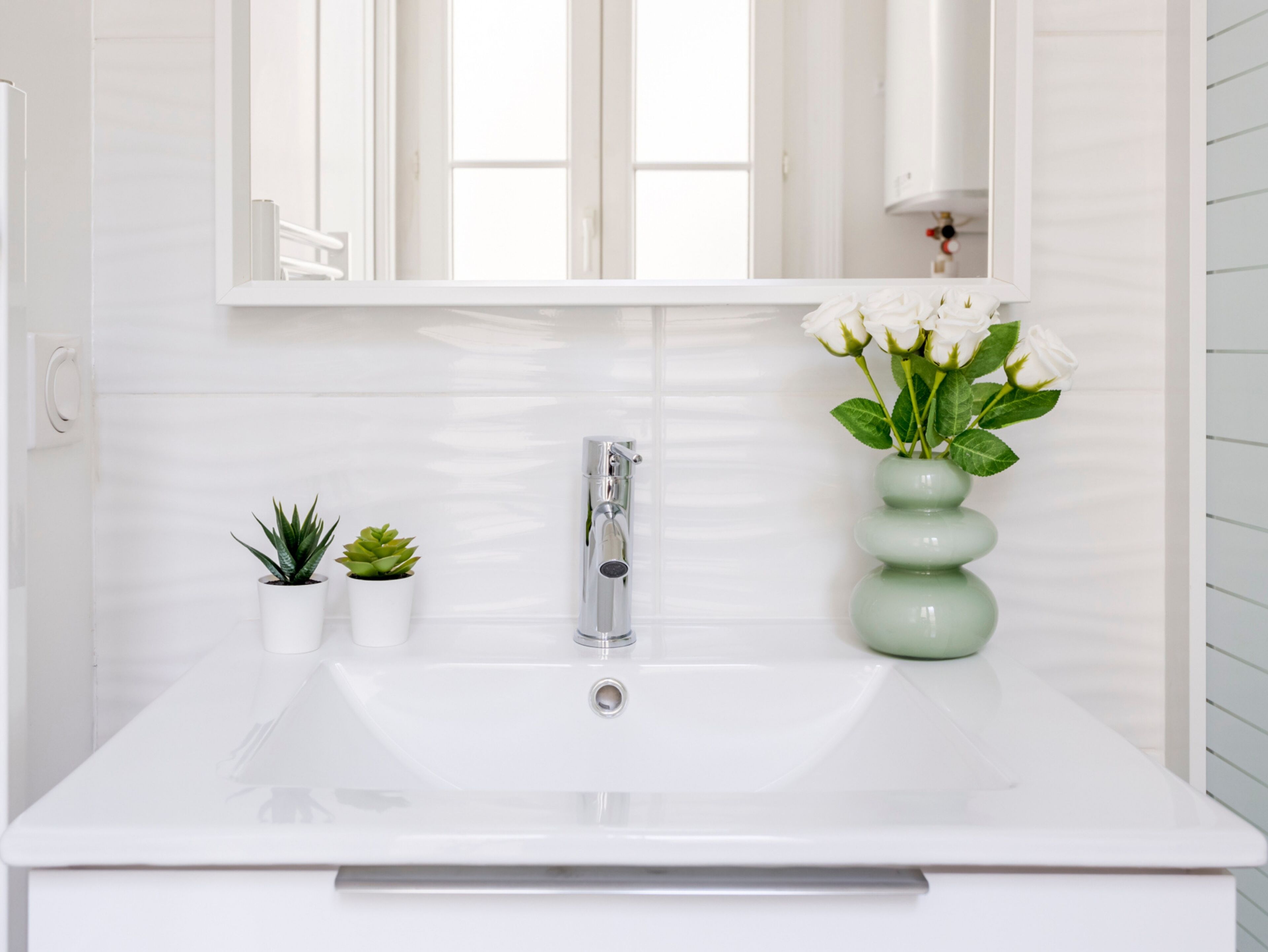 A white bathroom sink with a modern faucet, two small potted plants, and a vase with white flowers.