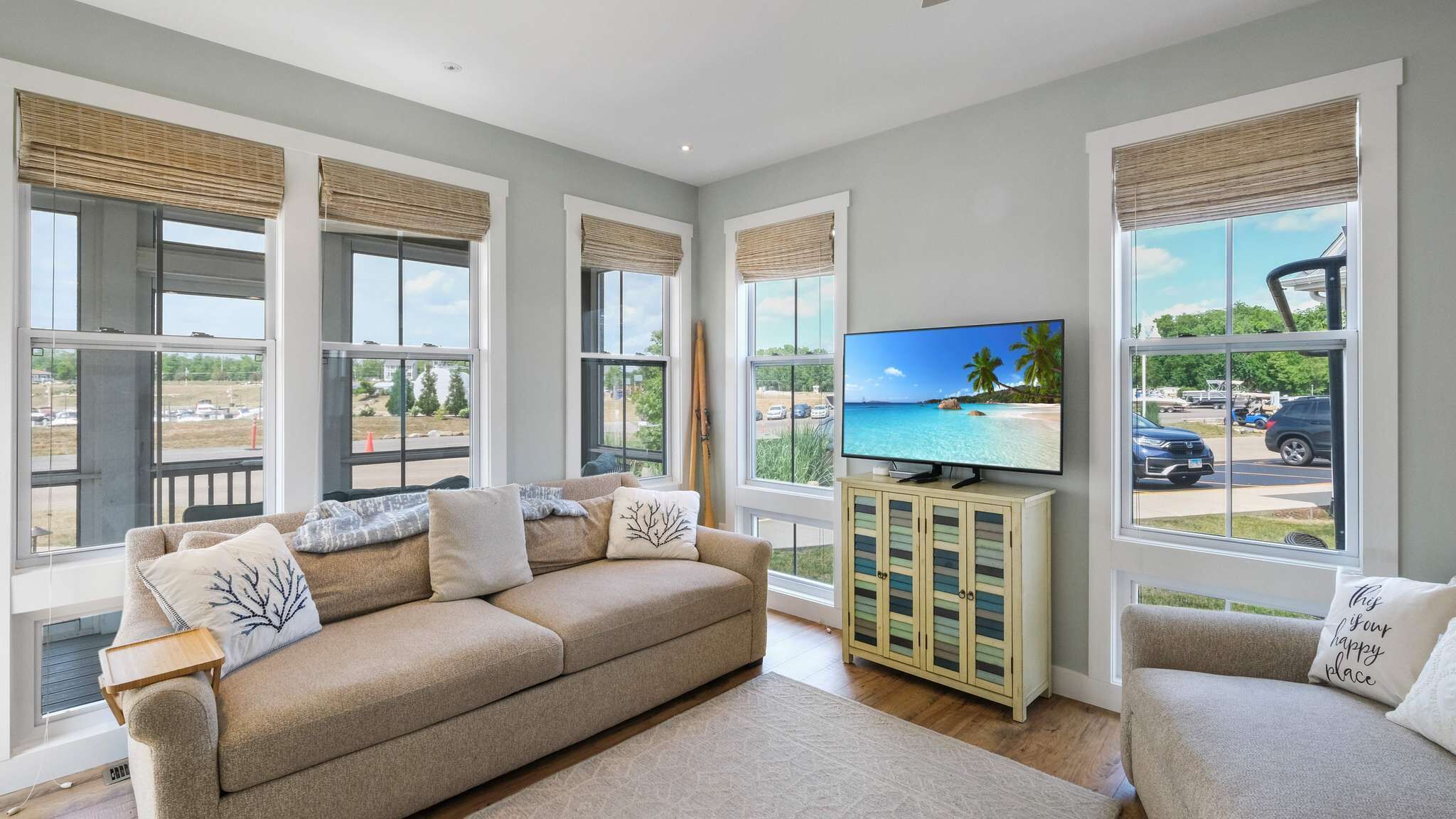 A living room with a beige sofa, a TV on a wooden stand, and large windows with blinds.