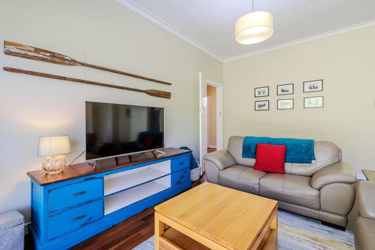 A living room with a TV on a wooden stand, a blue dresser, a beige sofa with a red pillow, and a wooden coffee table.