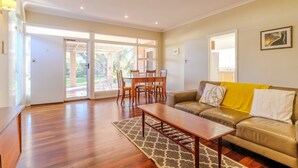 A spacious living room with a wooden floor, a yellow sofa, a dining table with chairs, and a sliding glass door leading to a patio. - Emu Point Beach Cottages (Emu Point)