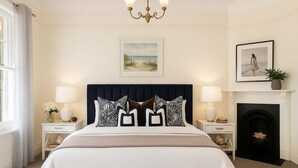 Une chambre à coucher avec un grand lit, deux tables de chevet, un lustre, une cheminée et des œuvres d’art accrochées aux murs.