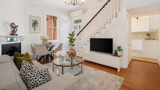 Un salon moderne avec un canapé, une table basse et un téléviseur. Un escalier mène à la cuisine.