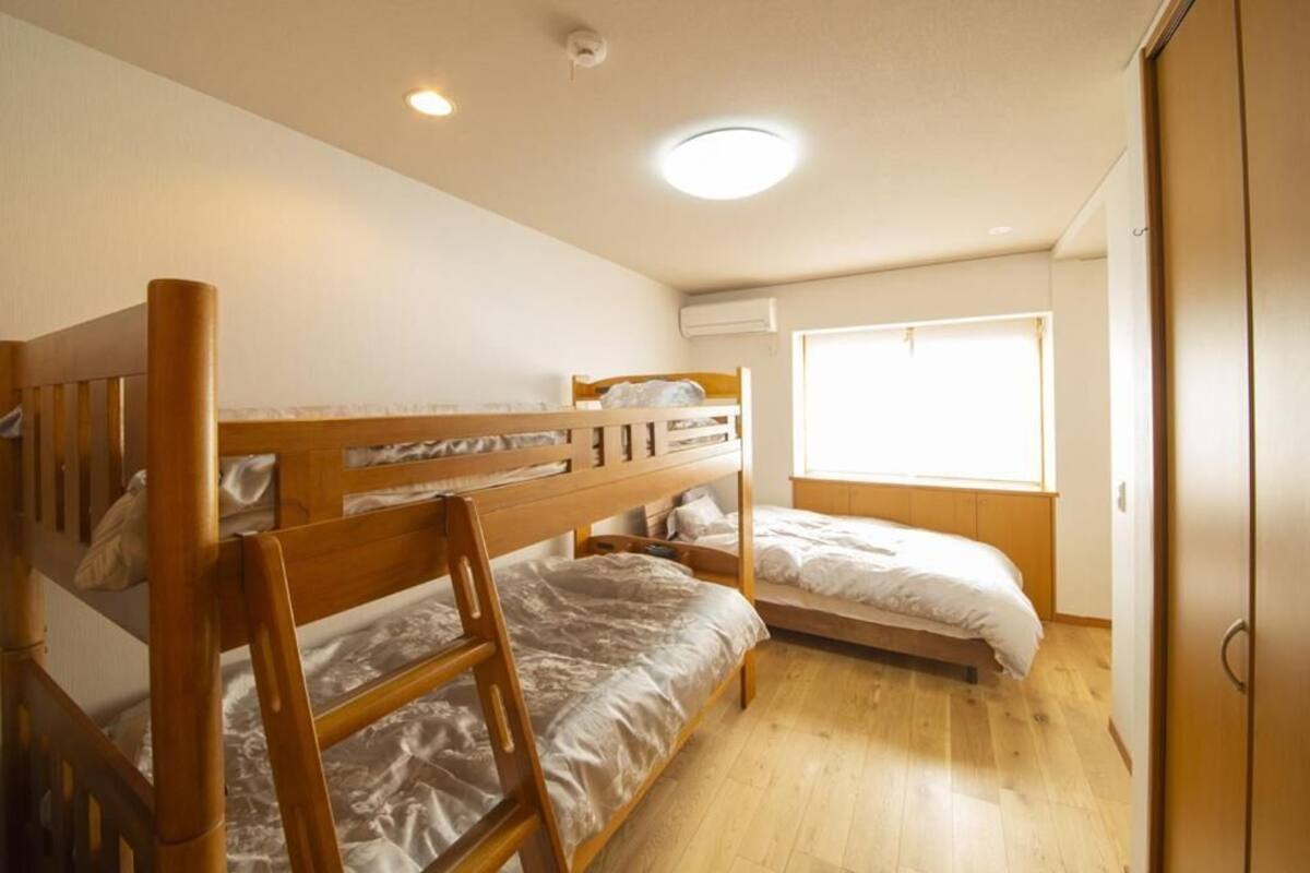 A dormitory room with two bunk beds, a wooden floor, a window with blinds, and a ceiling light.