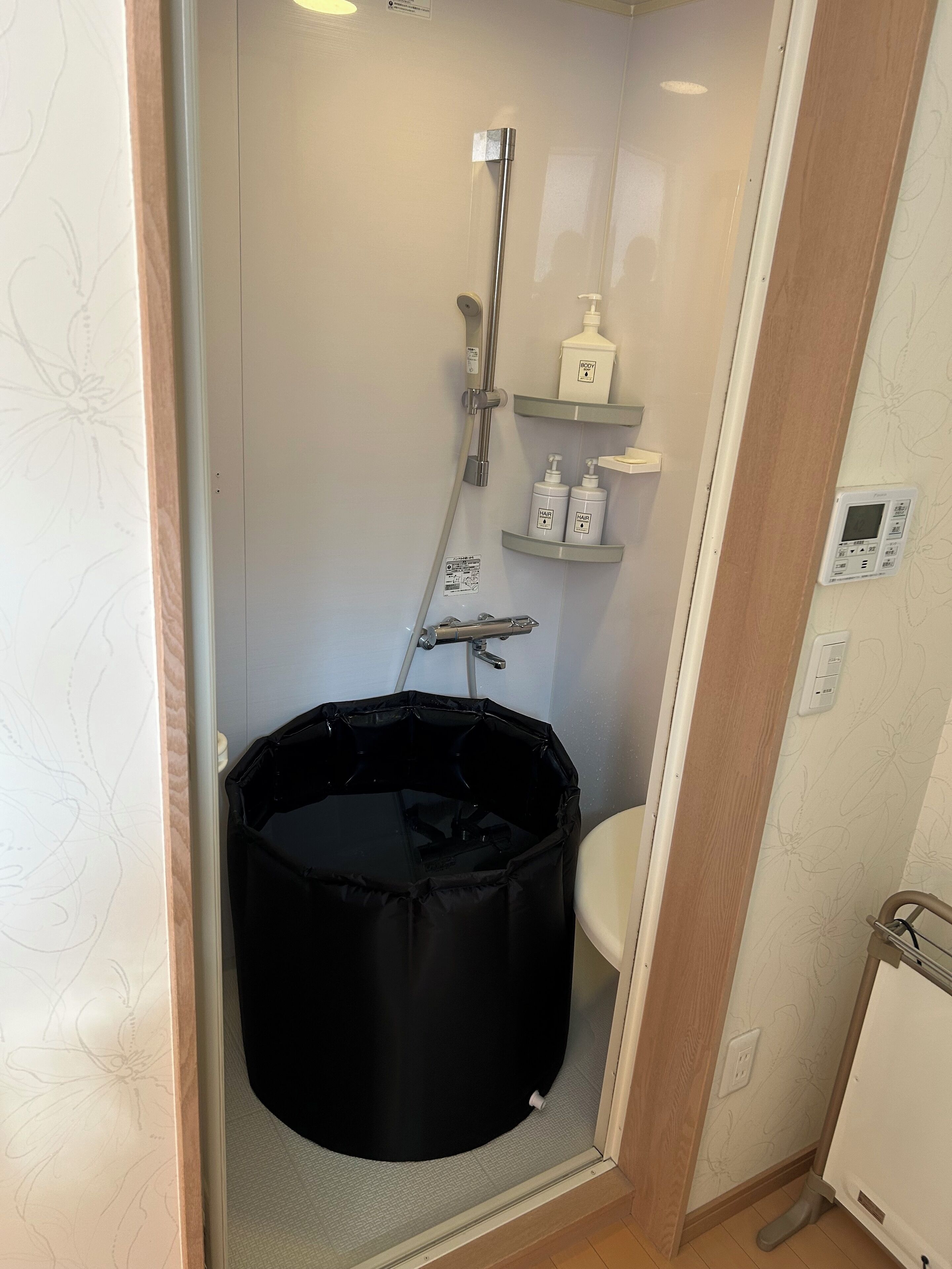 Casa de banho pequena com chuveiro, sanita e um recipiente de armazenamento preto.