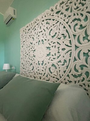 Een kamer met een decoratief wit hoofdbord, een bed met groene beddengoed, een witte lamp en een airconditioningunit aan de muur.