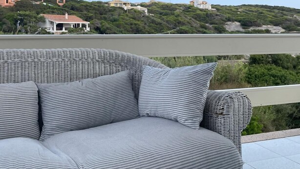 A wicker sofa with striped cushions on an outdoor balcony overlooking a body of water and a hilly landscape with buildings.