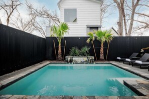 Une piscine rectangulaire bordée de pierres, entourée d’une clôture noire, de palmiers et de chaises longues.