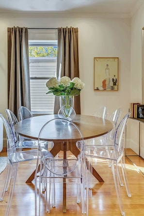 Uma sala de jantar com uma mesa redonda, cadeiras transparentes, um vaso com flores, uma janela com cortinas e um quadro pendurado na parede.