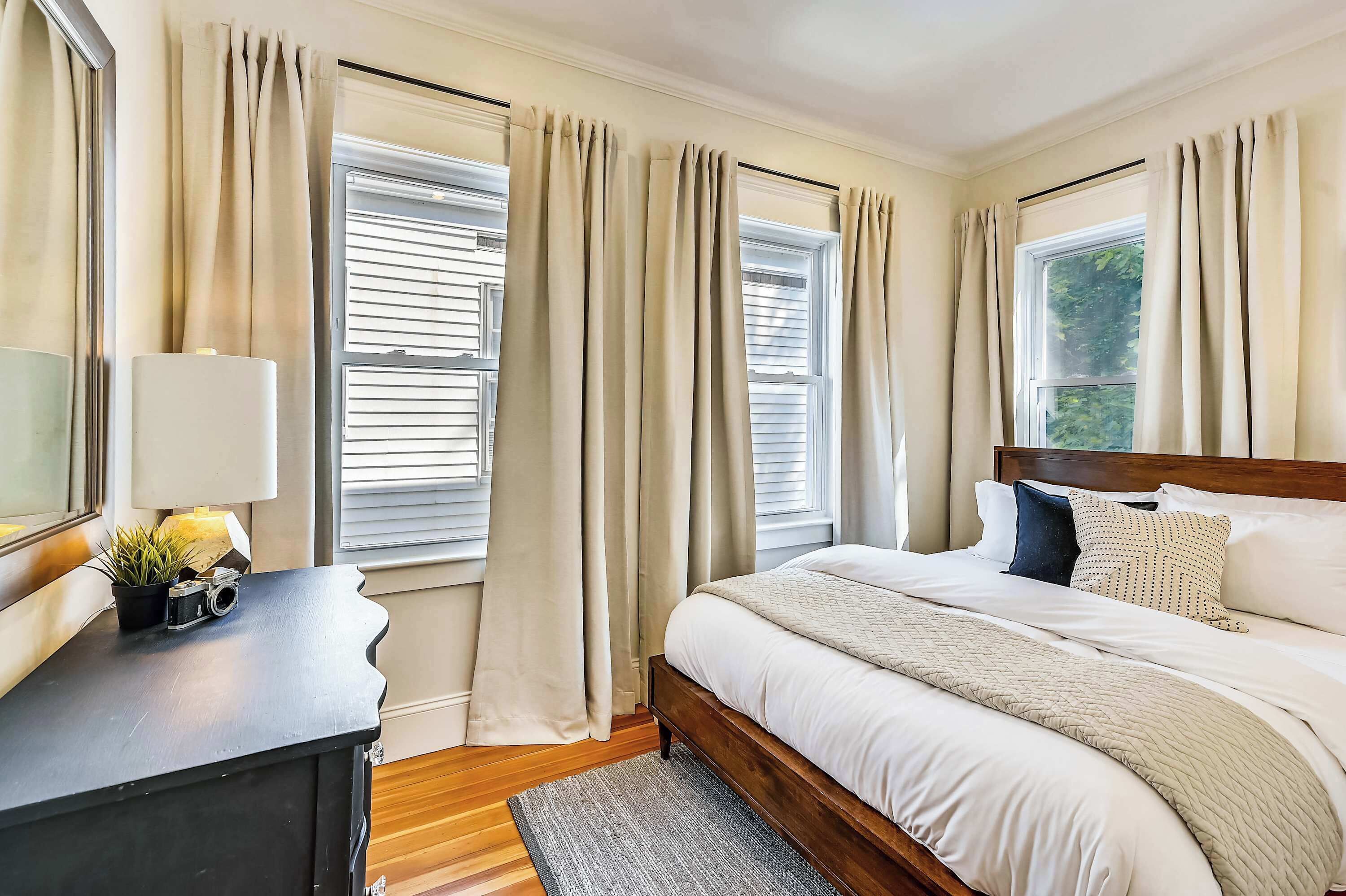 A bedroom with a bed, curtains, a lamp, and a small table with a plant and a camera.