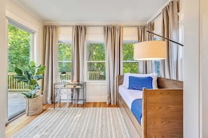 Un dormitorio con cama, escritorio, lámpara, ventana y vistas al verdor.