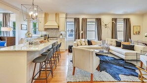 A modern kitchen with a central island, a dining area with bar stools, a living room with a sofa and a large rug, and a dining table with chairs. - 15BR Boston Entire Building | 3 Living Spaces (Boston)