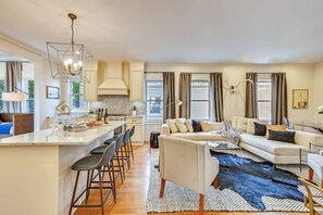 A modern kitchen with a central island, a dining area with bar stools, a living room with a sofa and a large rug, and a dining table with chairs. - 15BR Boston Entire Building | 3 Living Spaces (Boston)