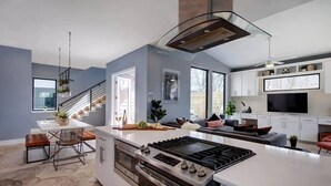 A modern kitchen with a central island, built-in oven, and a large stove. There is a dining area with a bench and chairs, and a staircase leading to another level.
