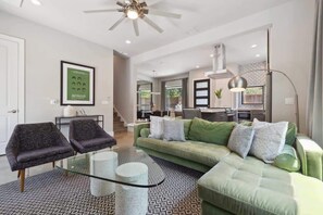 Un salon moderne avec un canapé vert, deux fauteuils, une table basse en verre et un tapis à motifs.