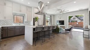 A modern kitchen with a central island, bar stools, and a dining area with a TV. - Luxe 5BR East Austin Home | Private Pool & Balcony (Austin)